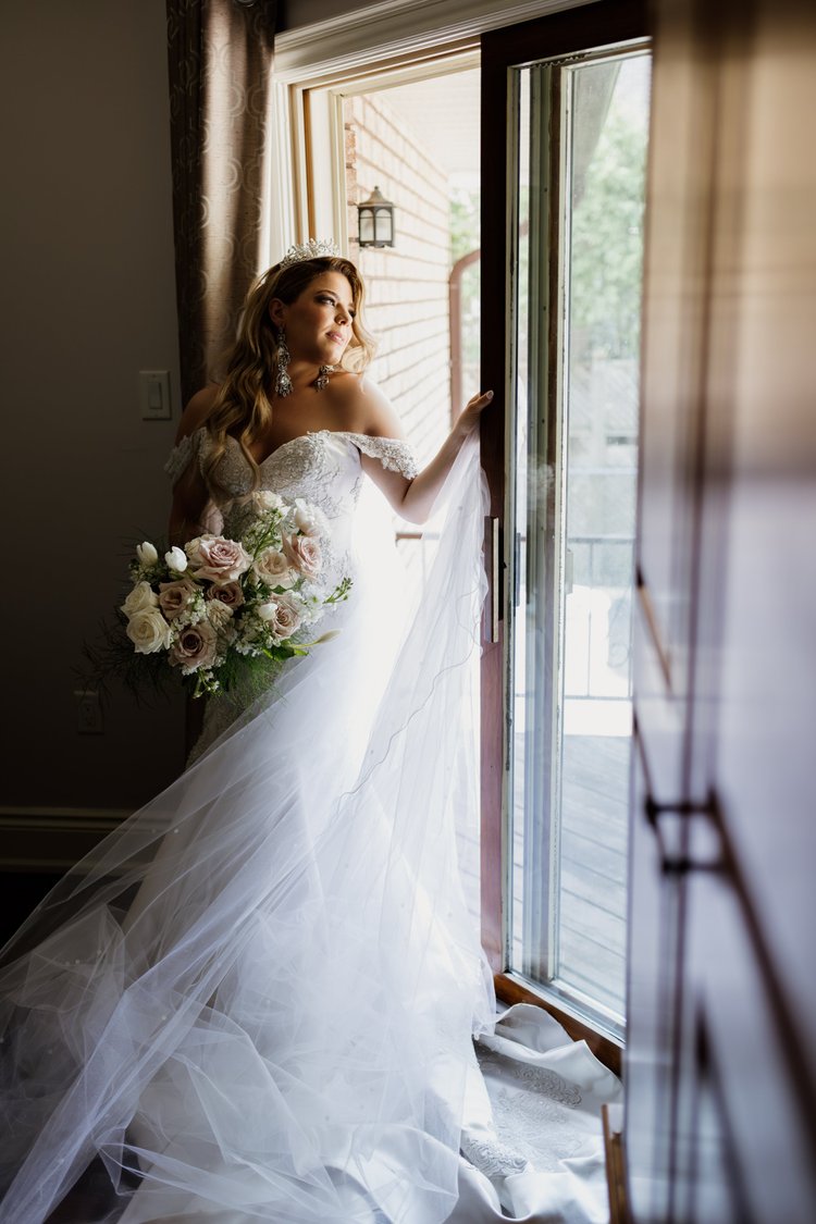Bride in a white wedding dress holding a bouquet, standing by a doorway, looking out.