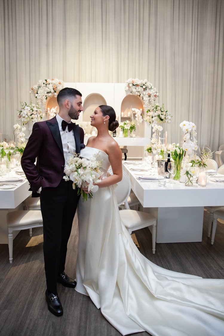 A bride and groom stand close together on their wedding day in a decorated reception room.