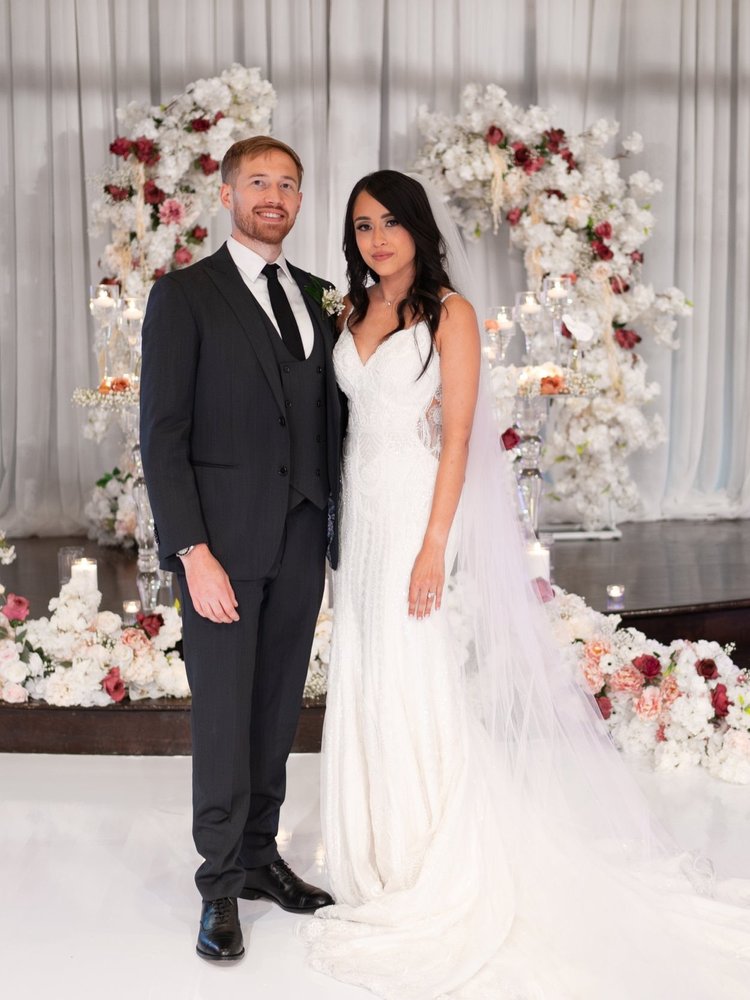 A bride and groom standing together in wedding attire in front of a floral backdrop.